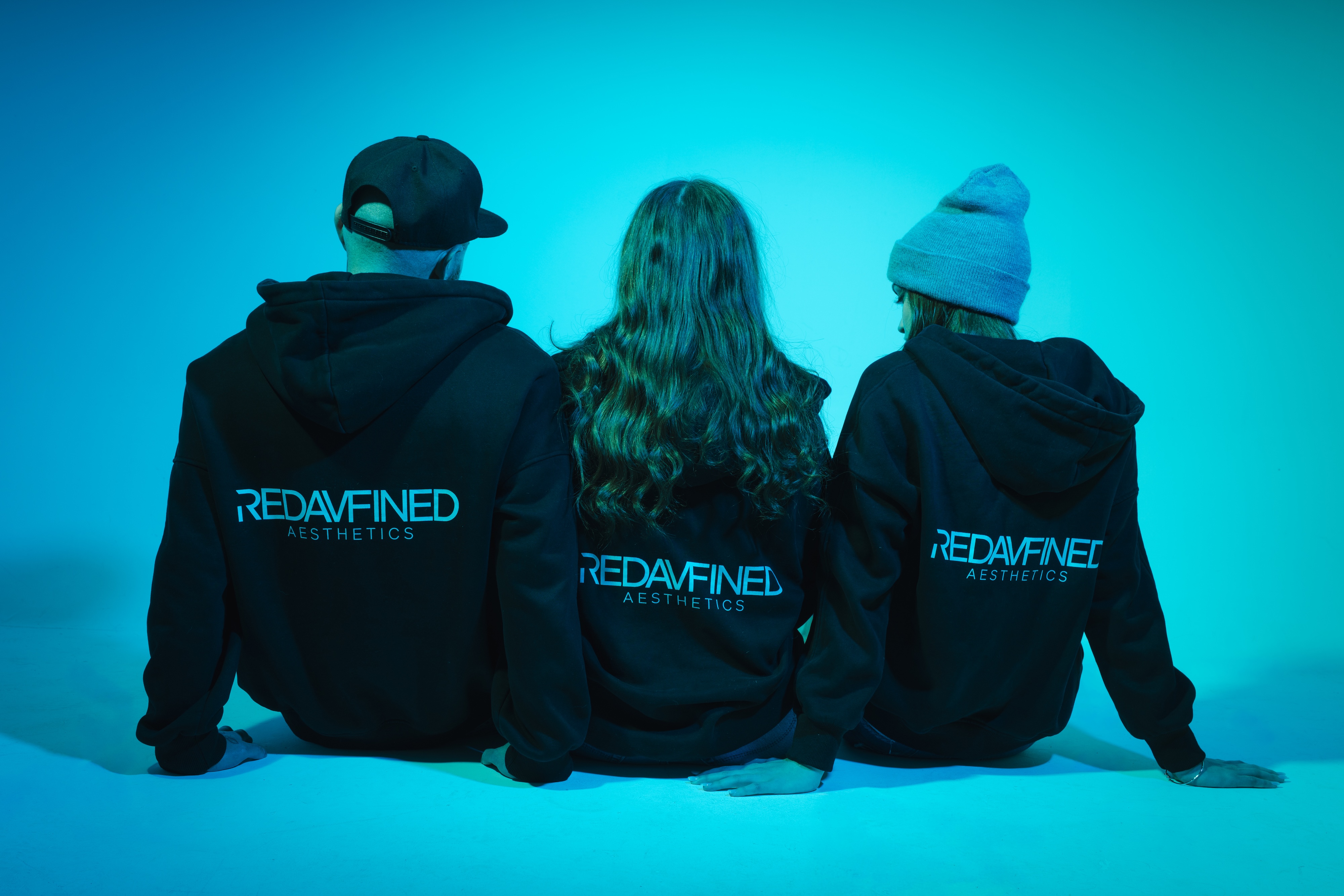 Three people in black Redavfined Aesthetics hoodies, seen from behind, against a blue backdrop
