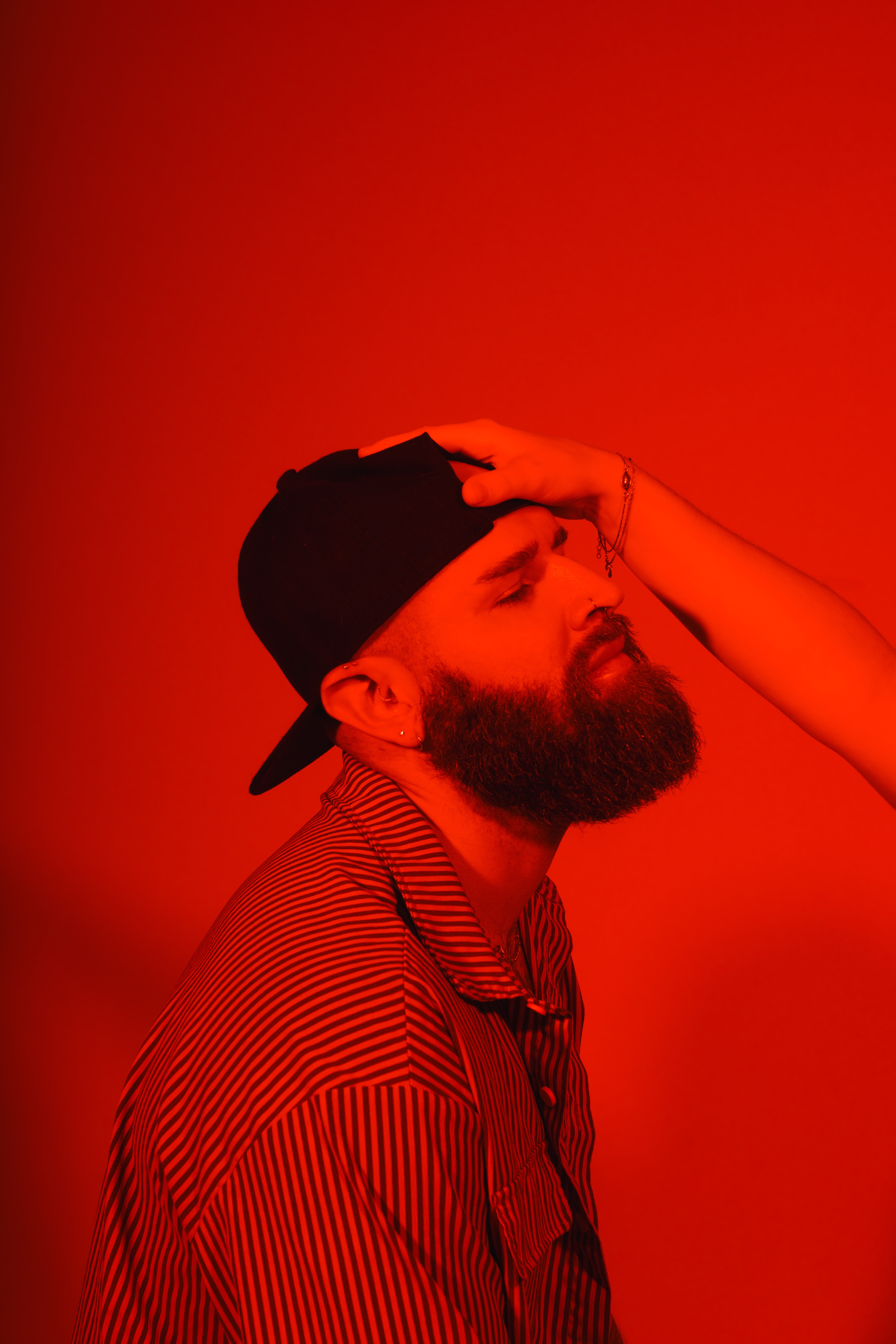 Portrait of a bearded man in a cap and striped shirt, eyes closed, against a red backdrop